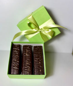 3 chocolate caramel bars laying in light green, square, paper box with a satin light green ribbon and bow wrapping the box.