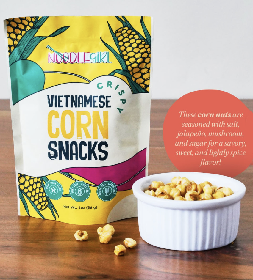 NoodleGirl Vietnamese Corn Snacks in a yellow and white stand-up pouch, shown next to a small white ramekin filled with corn nuts on a wooden surface. A red circular text bubble provides flavor and seasoning details.