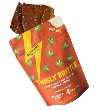 Brittle with green peppercorns spilling out of standup pouch packaging. Pouch is co branded with 50Hertz and NoCocoa with brown background and yellow and white text 