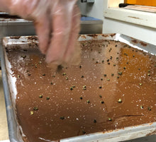 Load image into Gallery viewer, A slab of the toffee brittle in production. Gloved hand sprinkling sea salt and peppercorns on to melted chocolate in sheet pan