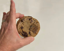 Load image into Gallery viewer, Hand holding a chocolate cookie with black sesame seed toffee brittle bits against a plain background