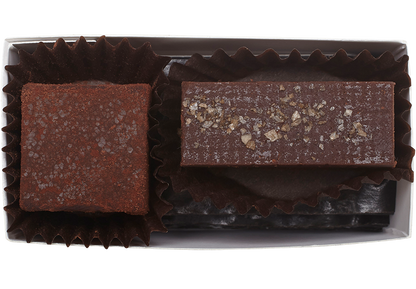 Above view and inside the rectangle box, one cube shaped Zested Lime truffle (coated in cocoa powder) and one rectangle shaped almond butter truffle with a few flakes of salt. Each truffle is in a brown paper candy cup. 